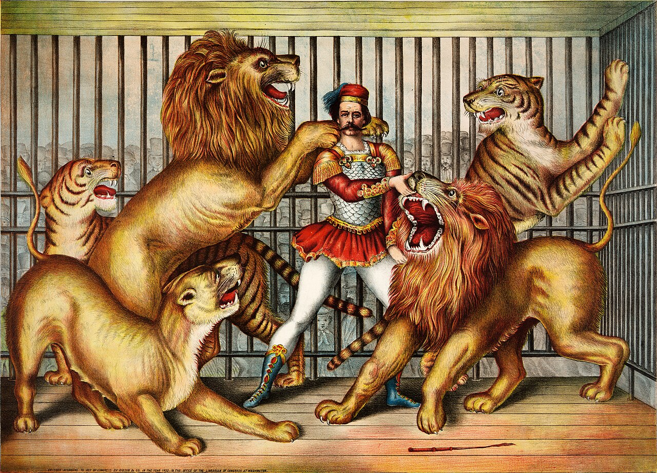 A colorful 19th-century chromolithograph of a lion tamer standing calmly inside a circus cage, surrounded by two lions, a lioness, and two tigers posed on pedestals around him.