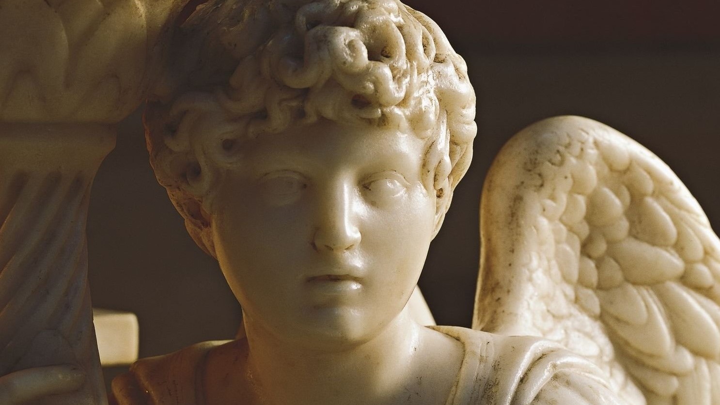 Close-up of a marble angel statue by Michelangelo Buonarroti with a calm, expressionless face and curly hair, warmly lit, with one textured wing visible behind its shoulder.